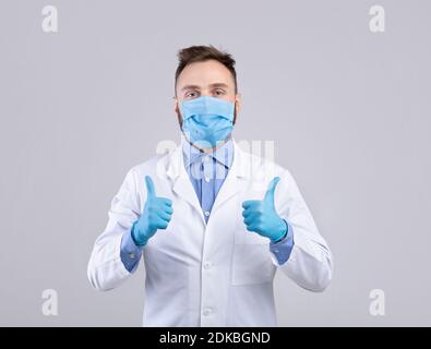 Millennial medico in uniforme, maschera viso e guanti guardando la macchina fotografica e mostrando i pollici verso l'alto gesto su sfondo grigio Foto Stock