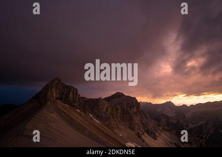 Il Breitgrieskarspitze con la sua suggestiva cresta occidentale nei Monti Karwendel, dopo il tramonto con un'atmosfera nuvolosa. Il paesaggio assomiglia ad un paesaggio lunare se non fosse per i resti della neve. Dal lato tirolese, l'umore oscuro di un temporale in avvicinamento. Foto Stock