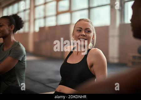 Giovane donna sorridente che parla con un gruppo diverso di amici mentre si siede su un pavimento della palestra dopo un allenamento Foto Stock