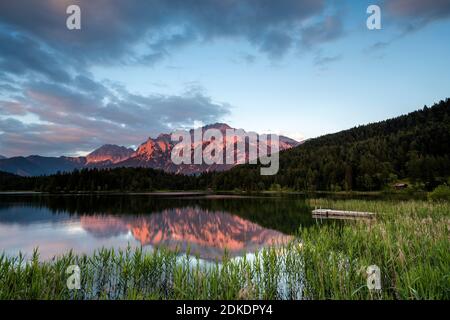 Il Lautersee vicino a Mittenwald riflette il bagliore alpino del Karwendel, con canne e un ponte pedonale in primo piano. Foto Stock