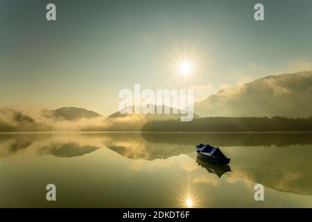 Al lago Sylvensteinspeicher è ormeggiato un'unica barca a remi con teloni, mentre il sole trasmette i suoi raggi sul lago con nebbia chiara al mattino presto e si riflette nell'acqua. Foto Stock