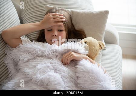 Little sick girl lies under the blanket and checks for fever on her forehead with her hand. Stock Photo