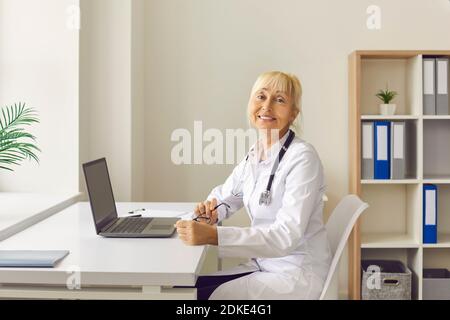 Positivo sicuro anziano donna dottore thetapist in uniforme medica bianca seduto vicino al computer portatile Foto Stock