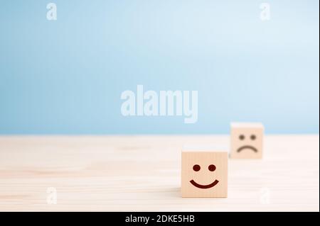 cubetti di legno con emoticon sorridenti e triste su tavolo di legno, sfondo blu. Spazio per il testo. Concetto di stato emotivo Foto Stock