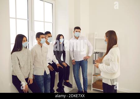 Giovani lavoratori in maschere di faccia ascoltando manager o allenatore In una riunione durante la pandemia di Covid-19 Foto Stock