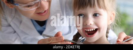 Donna dentista in occhiali protettivi esamina i denti della bambina paziente con strumenti metallici verticale Foto Stock
