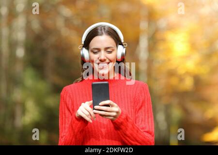 Vista frontale di una donna felice in rosso che ascolta musica con le cuffie wireless in un parco in autunno Foto Stock