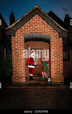 Regno Unito, Quorn - Dicembre 2020: Santa e un Elf si sbirciano fino a una porta d'ingresso mentre consegnano i regali Foto Stock