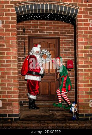 Regno Unito, Quorn - Dicembre 2020: Santa e un Elf si sbirciano fino a una porta d'ingresso mentre consegnano i regali Foto Stock