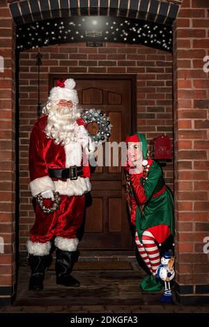 Regno Unito, Quorn - Dicembre 2020: Santa e un Elf si sbirciano fino a una porta d'ingresso mentre consegnano i regali Foto Stock