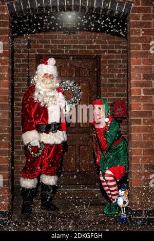 Regno Unito, Quorn - Dicembre 2020: Santa e un Elf si sbirciano fino a una porta d'ingresso, nella neve, mentre consegnano i regali Foto Stock