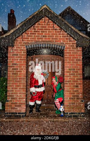 Regno Unito, Quorn - Dicembre 2020: Santa e un Elf si sbirciano fino a una porta d'ingresso, nella neve, mentre consegnano i regali Foto Stock