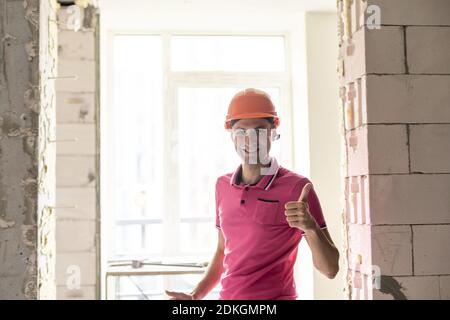 professione, costruzione e costruzione - felice lavoratore sorridente o costruttore in caschetto sopra stanza con attrezzatura di lavoro a casa nuova o appartamento sfondo Foto Stock