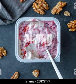 Yogurt bianco in tazza quadrata di plastica con marmellata e cucchiaio con noci su fondo grigio. Vista piatta. Foto Stock