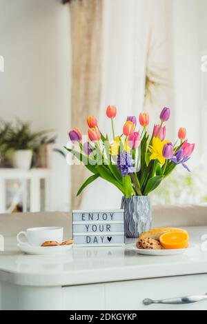 Concetto di Buongiorno. Colazione romantica - fiori freschi, tazza di bevanda calda, biscotti, arancio, scatola luminosa con messaggio Godetevi la vostra giornata sul tavolo in marmo con Foto Stock