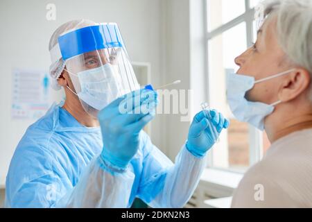 Lavoratore medico professionista che indossa dispositivi di protezione personale che testano la donna anziana per malattie pericolose utilizzando il bastone di prova Foto Stock