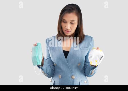 Donna asiatica di affari esita nella scelta di una maschera di salute sopra lo sfondo grigio Foto Stock