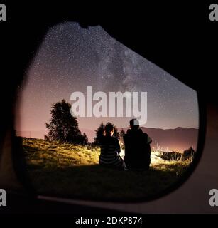 Vista posteriore di felici e romantici coppie che riposano al campeggio accanto al falò sotto il cielo notturno incredibile pieno di stelle e Via Lattea. Sullo sfondo cielo stellato, montagne. Vista dall'interno tenda turistica Foto Stock