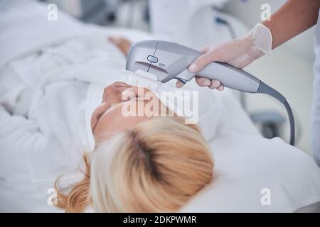 Bella donna bionda sdraiata sul lettino elettrico di bellezza mentre riceve cura della pelle del viso Foto Stock