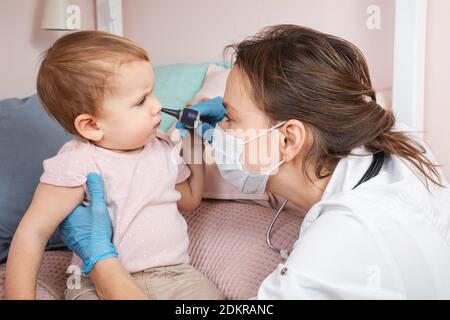 Il pediatra esamina il naso della bambina a casa durante la quarantena pandemica del coronavirus COVID-19. Medico che usa otoscopio (auriscopio) per controllare il passaggio nasale Foto Stock