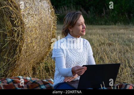 bella ragazza giovane in un maglione bianco sullo sfondo di un haystack con un laptop in mano si siede a terra, guarda in lontananza Foto Stock