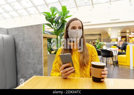 Bruna giovane donna felice in maschera protettiva con tazza di caffè seduto al tavolo in centro commerciale o caffè. Colori giallo e grigio. Foto Stock