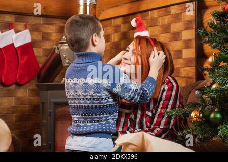 Un ragazzino mette un cappello di Natale sulla madre sullo sfondo di un albero di Natale decorato, un camino illuminato, calze appesi per regali .. Foto Stock