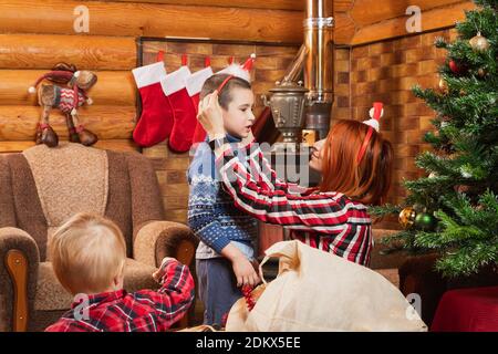 Mamma mette un cappello di Natale sul suo figlio piccolo, un albero di Natale decorato sullo sfondo, un camino illuminato, calze appesi per regali .. Buon Natale. Foto Stock