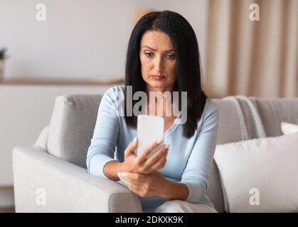 Donna seria matura che usa lo smartphone a casa sua Foto Stock