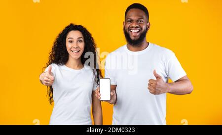 Coppia eccitata che mostra lo schermo del telefono gesturando Thumbs-Up su sfondo giallo Foto Stock