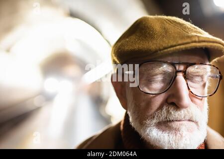 ritratto di uomo anziano in cappuccio e occhiali in piedi occhi chiusi sulla piattaforma della metropolitana con luce nel tunnel della metropolitana sullo sfondo Foto Stock