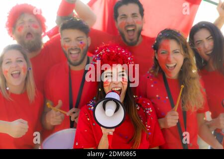 Tifosi di calcio urlando, con camicie rosse nello stadio. Gruppo di giovani molto entusiasti dello sport. Concetto di sport e divertimento. Mettere a fuoco sulla faccia della ragazza con Foto Stock