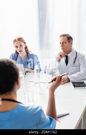 Medico e infermiera che guardano il collega afroamericano su offuscato in primo piano mentre si lavora vicino a dispositivi e carte Foto Stock