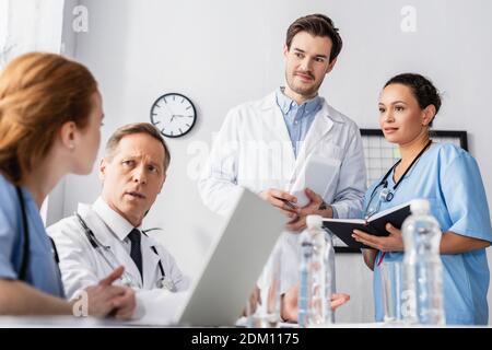 Personale ospedaliero multietnico con notebook e dispositivi che lavorano vicino all'acqua in primo piano sfocato sul tavolo Foto Stock