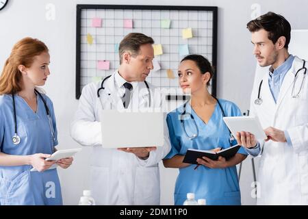 Personale ospedaliero multiculturale con dispositivi e notebook che lavorano in clinica Foto Stock