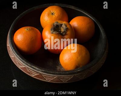 Un pezzo di frutta Kaki circondato da quattro arance una ciotola Foto Stock