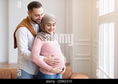 Coppia musulmana in attesa di un bambino, abbracciando in piedi a casa Foto Stock