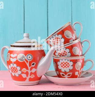 Una pila di tazze retro in ceramica con teiera a motivi rossi su un tavolo di legno rosa contro uno sfondo blu di legno, tendenza colore pastello Foto Stock