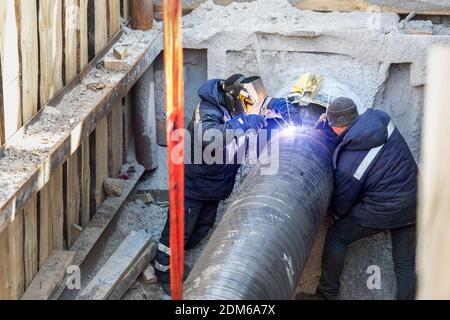 Professional welder welding water or gas steel pipeline in protective trench and mask at construction site pit. City underground utilitites renewal Stock Photo