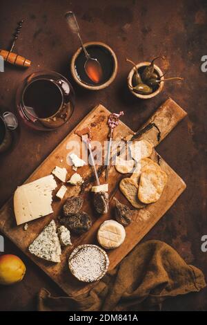 Spuntini e antipasti per un concetto di vino rosso. Piatto di vari formaggi, cracker, carne affumicata e bicchiere di vino rosso su tavola rustica di legno su sfondo marrone arrugginito, vista dall'alto Foto Stock