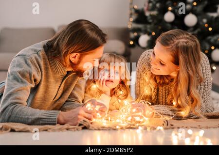 La famiglia felice con la figlia si trova sul pavimento e accogliente colloqui dalla luce delle ghirlande sullo sfondo di Albero di Natale Foto Stock