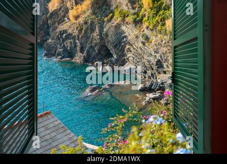 Vista da una finestra aperta su una piccola baia con nuotatori seduti sulle rocce, vicino al villaggio di Vernazza, parte delle cinque Terre Foto Stock