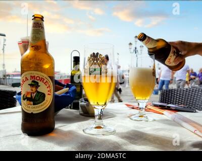 Una mano femminile versa una bottiglia di birra italiana Un bicchiere su un tavolo da patio all'aperto a Venezia Italia Foto Stock