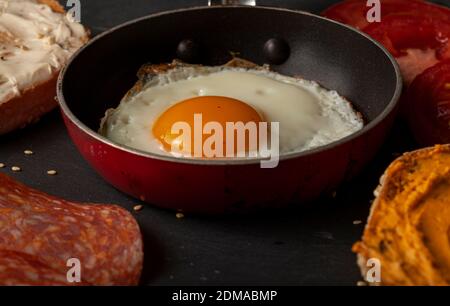 primo piano immagine macro di un uovo fritto (lato soleggiato in alto) in una padella antiaderente su un tavolo di legno nero. pomodori, toast macchiati e cremati e fette di sala Foto Stock