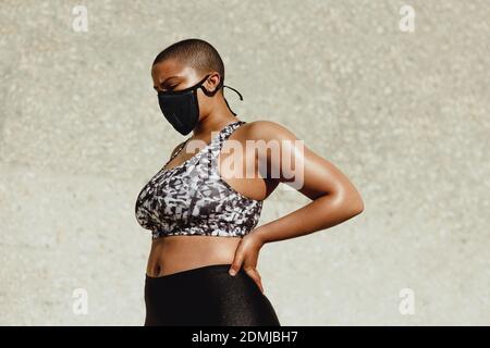 Donna che prende la pausa dopo l'allenamento all'aperto. Donna che indossa abiti sportivi e maschera protettiva per il viso che riposa dopo la sessione di allenamento. Foto Stock