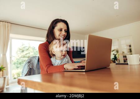 Madre sorridente e bambino seduto al tavolo e lavorando su un computer portatile. Donna con bambino che usa il laptop mentre lavora da casa. Foto Stock