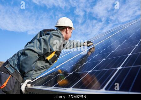 Vista laterale istantanea del workman, indossando uniforme, guanti da lavoro e casco, impostando una nuova batteria solare lucida con l'aiuto di chiave esagonale, cielo blu su sfondo. Concetto di energia verde Foto Stock