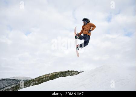 Guy snowboarder in giacca invernale e casco che salta in aria. Adorabile bambino che fa saltare con snowboard mentre scivola giù collina innevata nelle montagne invernali. Concetto di attività sportive invernali. Foto Stock