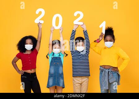 Carino misto razza bambini che indossano maschere mediche che tengono 2021 numeri isolati su sfondo giallo, nuovo anno nel tempo dei concetti pandemici Foto Stock