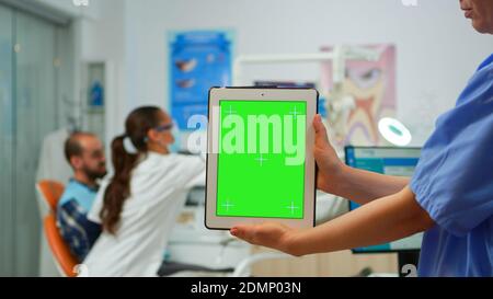 Primo piano della compressa con schermo verde tenuto da un assistente dentale in piedi nella stanza stomatologica, mentre il medico esamina il paziente in background. Utilizzo di monitor con tastiera pc con chiave chroma ziolata mockup Foto Stock